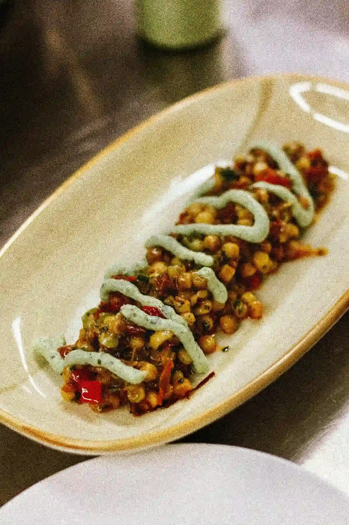 Länglicher Teller mit Kichererbsen, Mais, Paprika, grüner Soße auf Edelstahloberfläche.