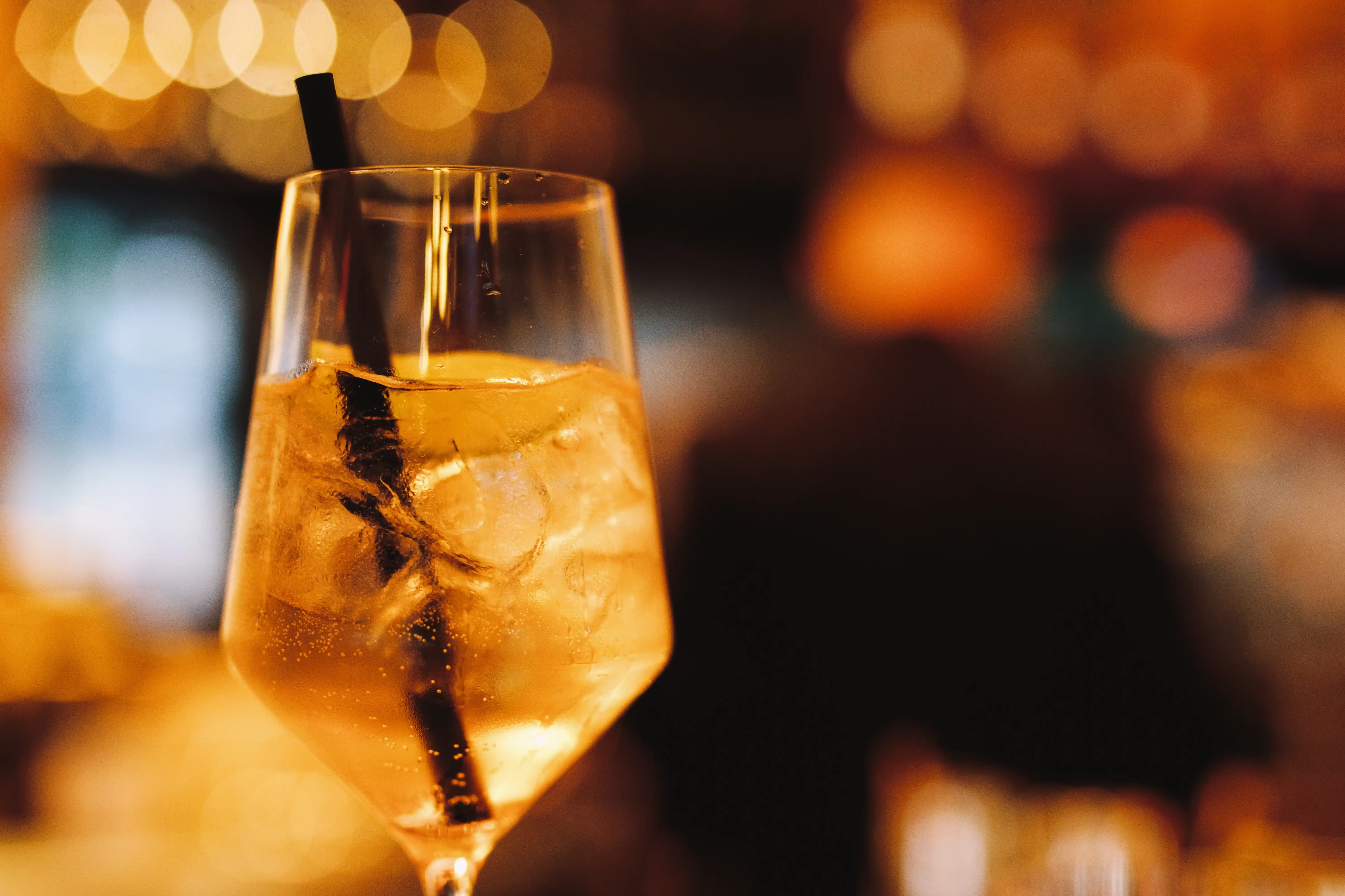 Ein Glas mit golden schimmerndem Aperitif und Eiswürfeln, mit schwarzem Strohhalm, vor unscharfem, warm beleuchtetem Hintergrund in einer Bar.