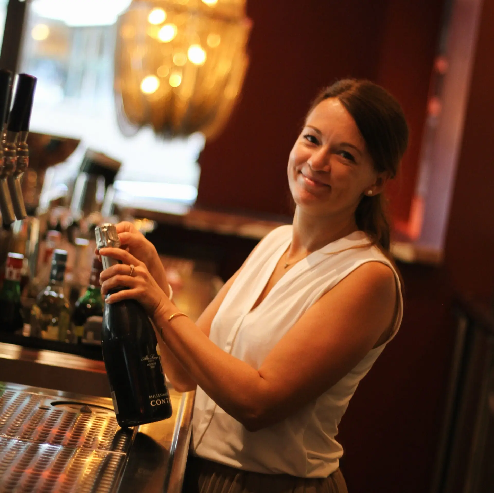 Lächelnde Frau öffnet eine Flasche Sekt an der Bar mit warmen Licht.